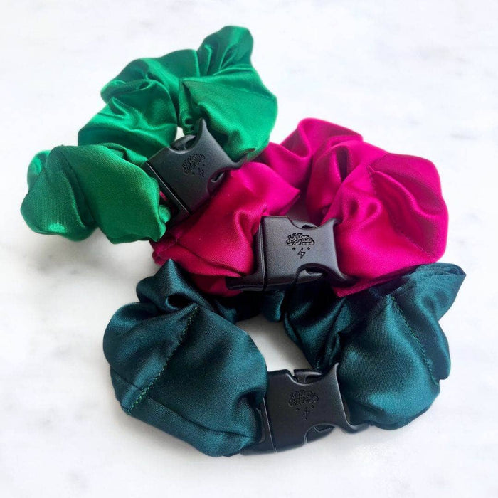 Three satin scrunchies in green, pink, and teal with black plastic clips on a white background.