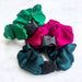 Three satin scrunchies in green, pink, and teal with black plastic clips on a white background.