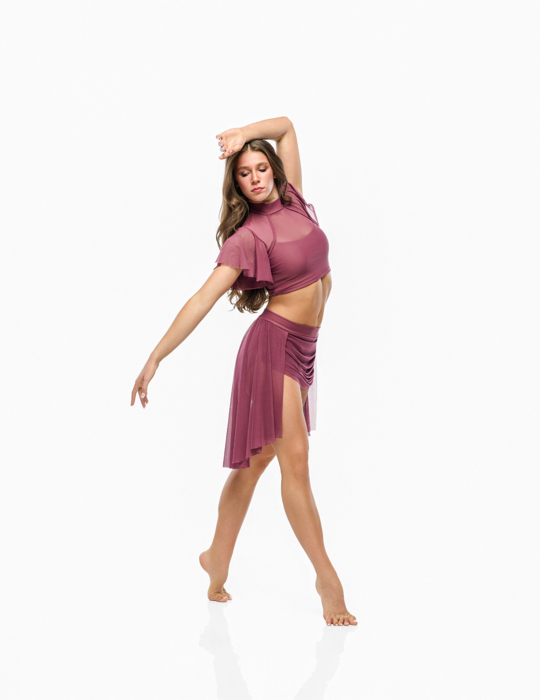 A woman modeling a dusty rose two-piece dance or yoga set consisting of a sheer short-sleeve crop top and a matching ruched wrap skirt, posed against a seamless white background.