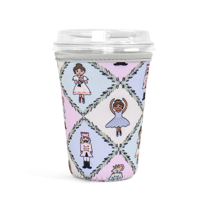 A reusable coffee cup with a clear lid and a pastel diamond-patterned sleeve featuring ballerina and nutcracker illustrations, shown on a white background.