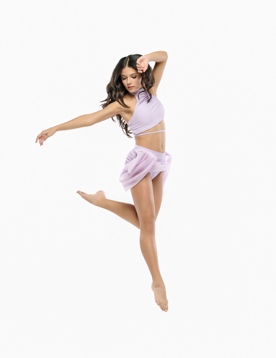 A woman in a light lavender two-piece dance or yoga outfit is captured mid-jump against a solid white background. The outfit consists of a halter-style crop top and a matching sheer ruffled mini skirt.