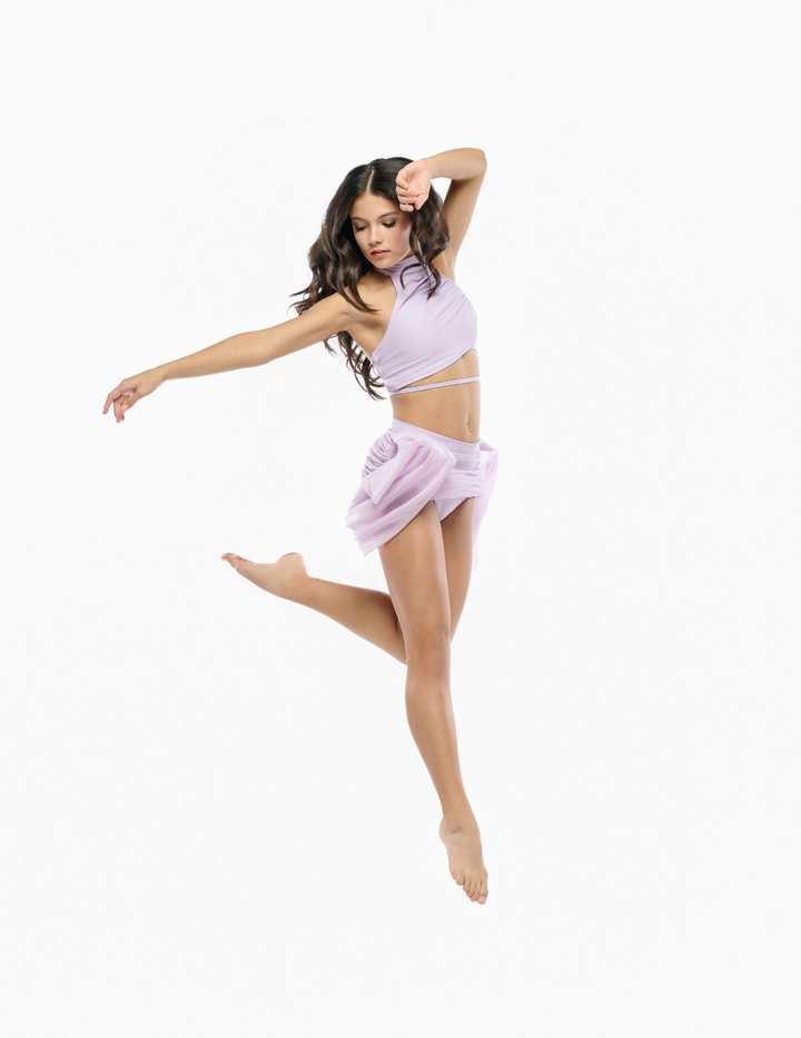 A woman in a light lavender two-piece dance or yoga outfit is captured mid-jump against a solid white background. The outfit consists of a halter-style crop top and a matching sheer ruffled mini skirt.