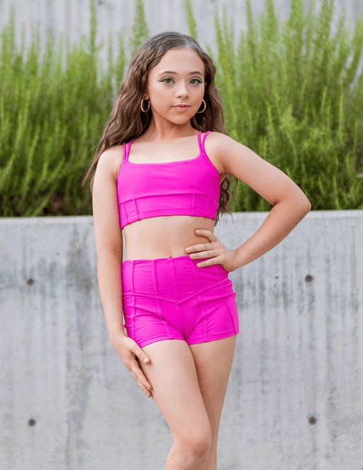 Young girl in bright pink athletic outfit standing outdoors with greenery in the background