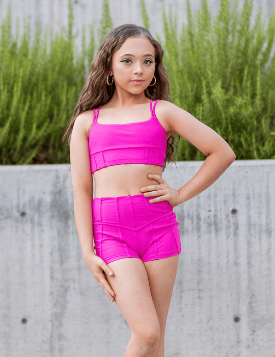 Young girl in bright pink athletic outfit standing outdoors with greenery in the background