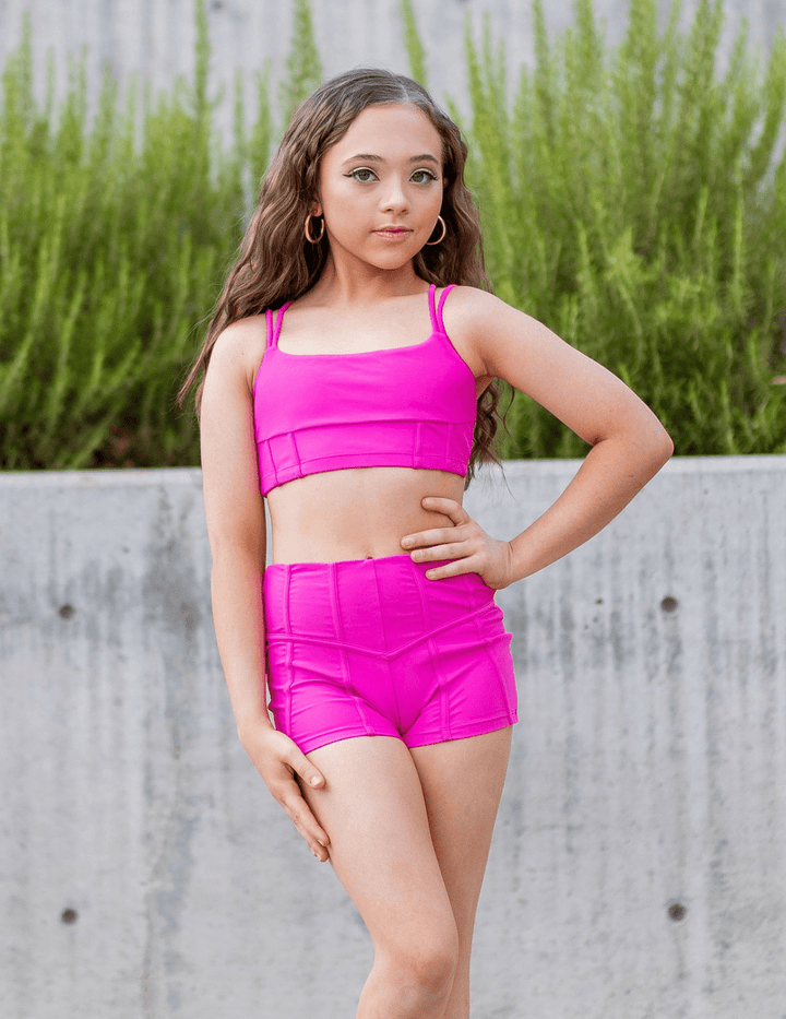 Young girl in bright pink athletic outfit standing outdoors with greenery in the background
