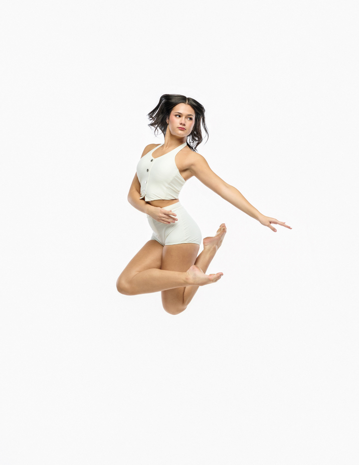 A woman in a white two-piece dance or gymnastics leotard jumps against a solid white background.
