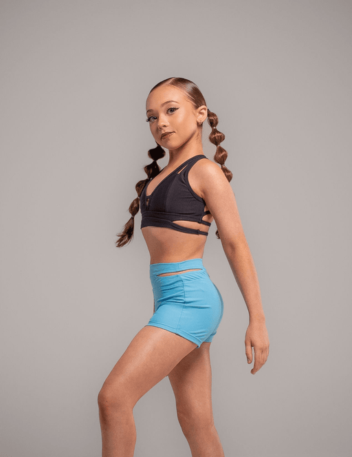 Young girl in black sports bra and blue shorts posing against a gray background