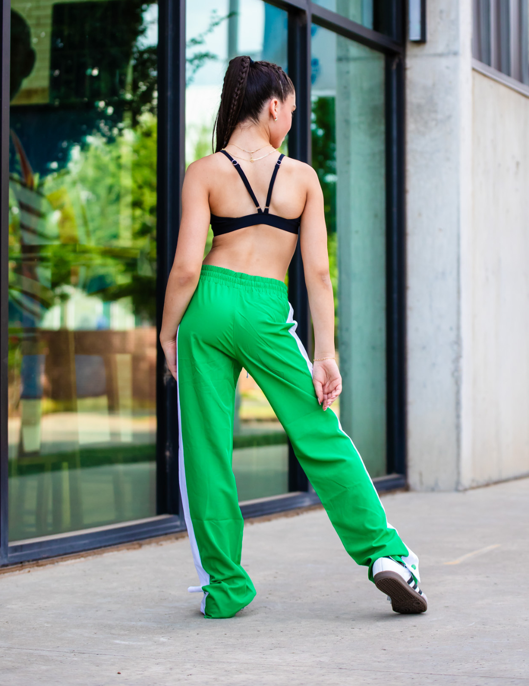 Woman wearing a black sports bra and green tracksuit with white stripes, standing in front of a glass building.