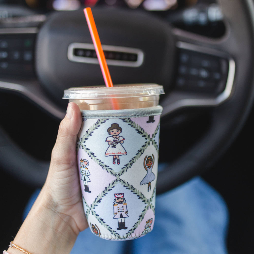 A hand holds a clear plastic travel tumbler with a pink neoprene sleeve featuring a vintage-style illustration of princesses and princes in a diamond pattern. The cup has a clear lid and an orange straw, with a blurred car steering wheel in the background.