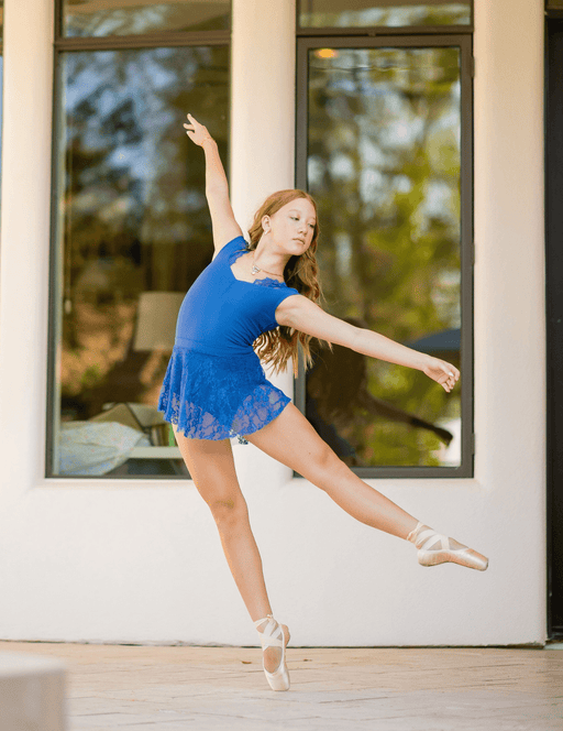 Dancer in a blue outfit practicing ballet outside a building with large windows.
