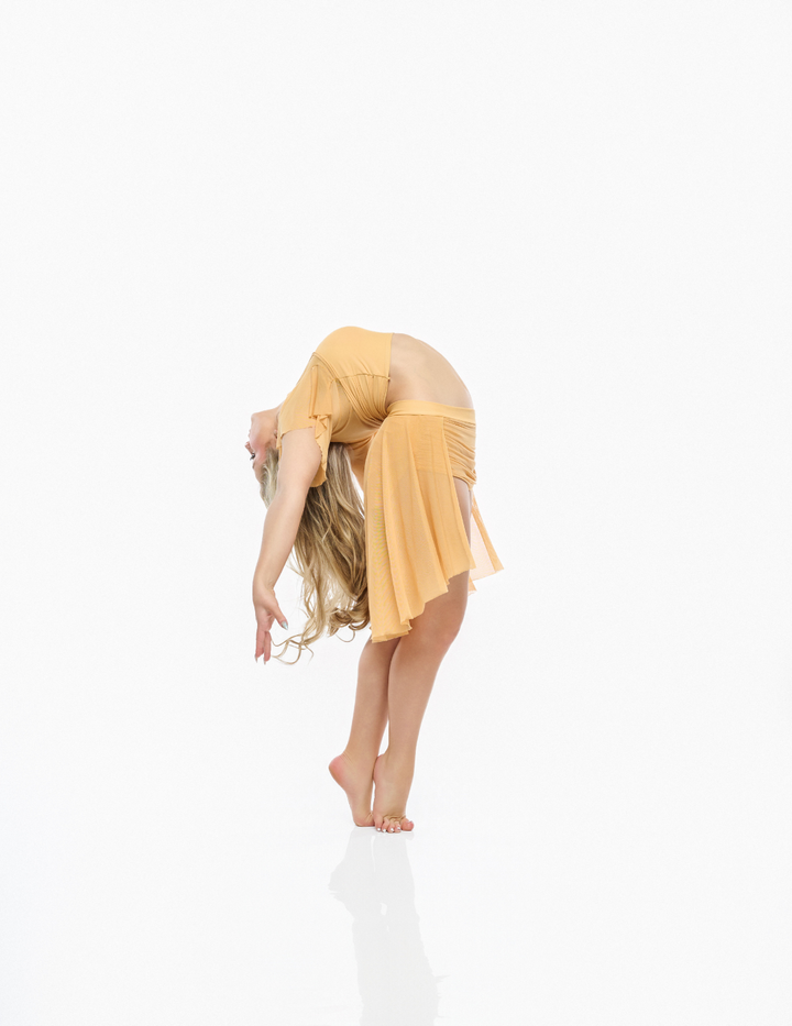 A woman in a flowing mustard yellow dance or yoga outfit bends forward on a reflective white studio floor against a solid white background.