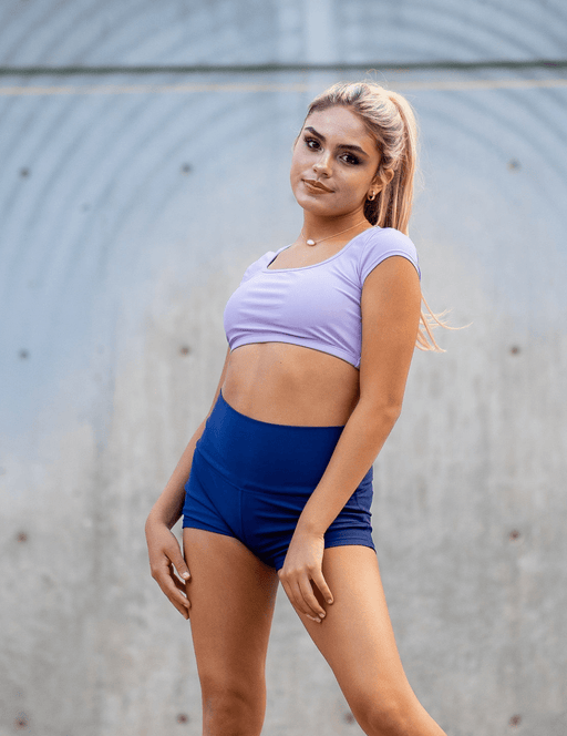 Woman wearing a purple crop top and blue shorts against a gray wall.