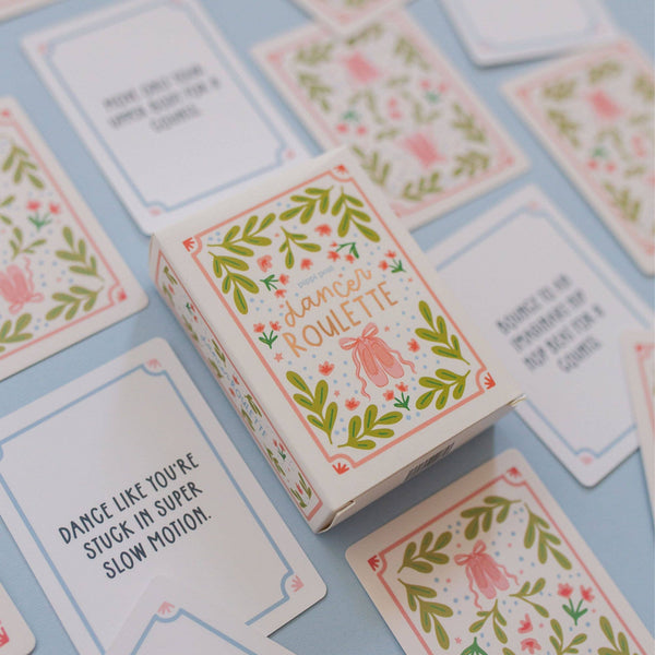 A boxed card game called "dance roulette" sits on a light blue surface with several illustrated cards scattered around it. The box is white with pink and green botanical border art and the title in peach lettering.