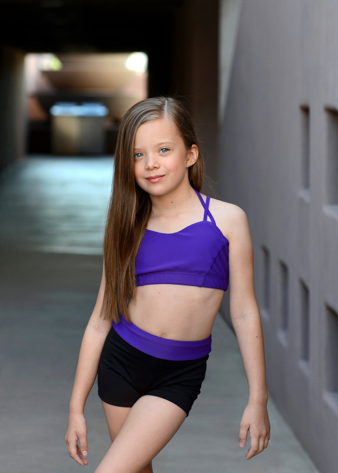 Young girl in a purple sports bra and black shorts standing against a neutral background