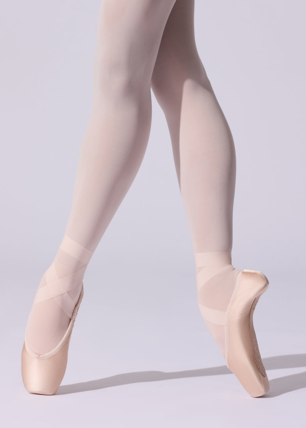 A close-up of a dancer's legs in nude tights and satin pointe shoes, posed en pointe against a light gray studio background.