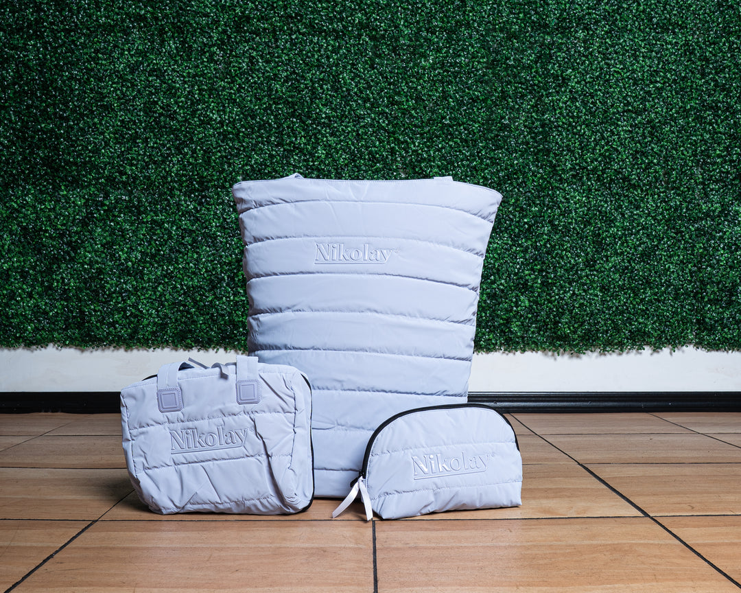 Lilac quilted bags with 'Nikolay' branding on a wooden floor against a green grass wall.