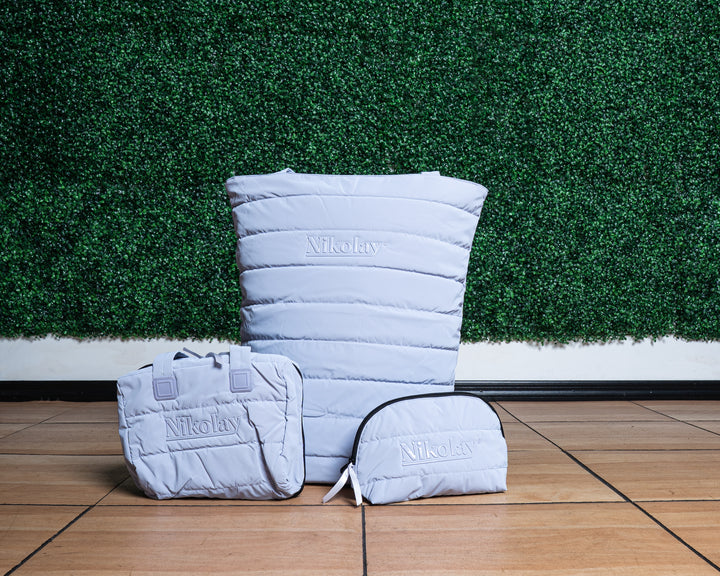 Lilac quilted bags with 'Nikolay' branding on a wooden floor against a green grass wall.