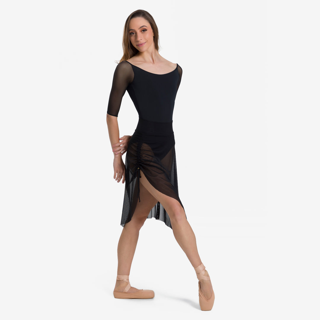 A woman modeling a black dance leotard with sheer three-quarter sleeves and a matching sheer wrap skirt, paired with nude ballet shoes, against a plain white studio background.