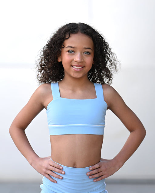 Young girl wearing a light blue sports bra and leggings against a plain background