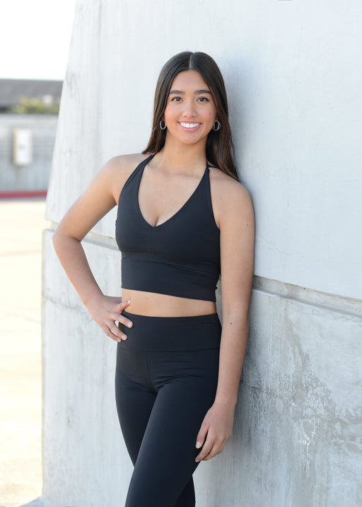 Woman wearing a black sports bra and leggings leaning against a white wall.