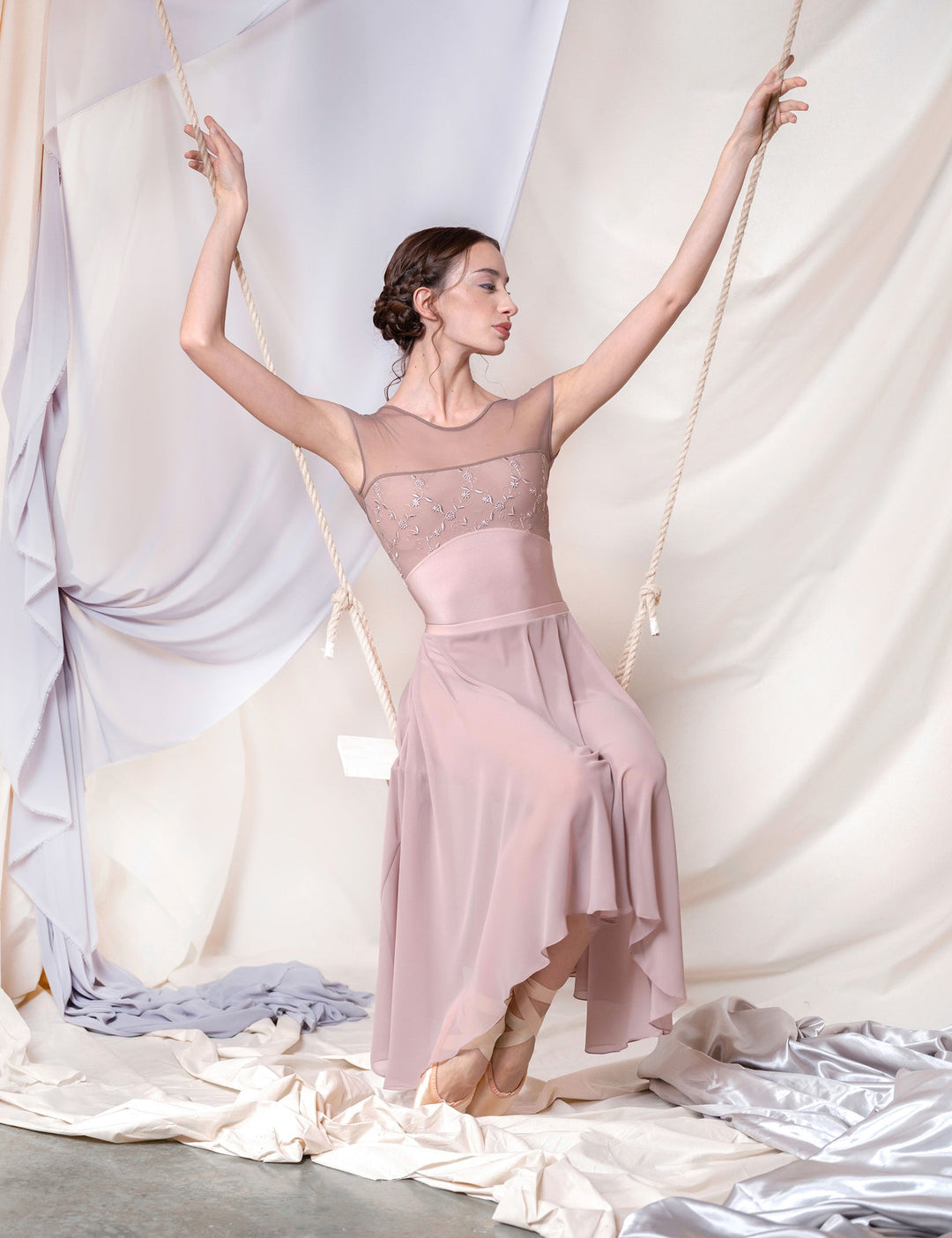 Ballerina in a pink dress posing gracefully with arms outstretched against draped fabric.