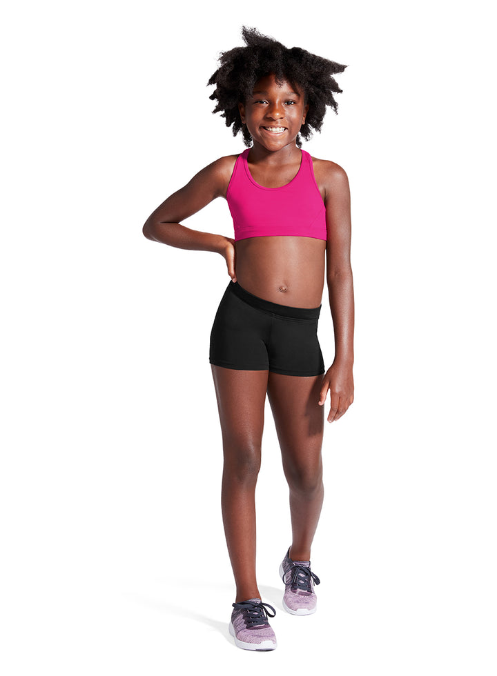 A young girl with curly black hair stands against a white background wearing a bright pink athletic sports bra and black athletic shorts, paired with gray and purple sneakers.