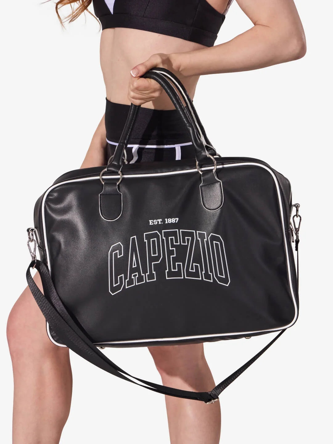 Person holding a black duffel bag with 'Capezio' branding on a white background