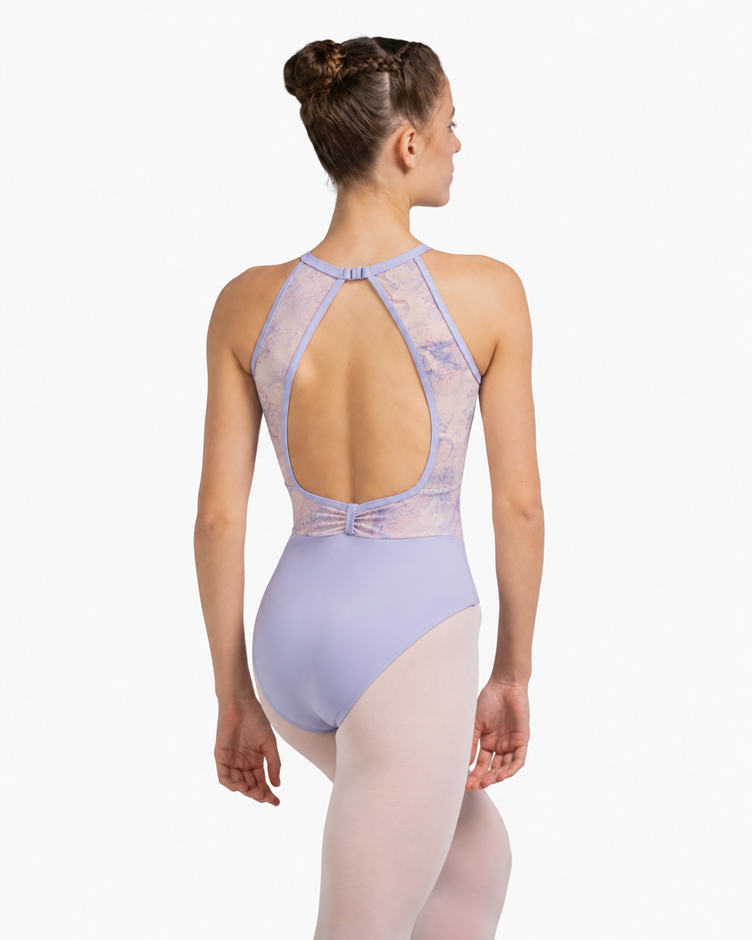 A woman modeling a lavender ballet leotard from the back, showing a high open back with sheer mesh straps and a bow detail at the waist, paired with light pink tights against a light gray background.