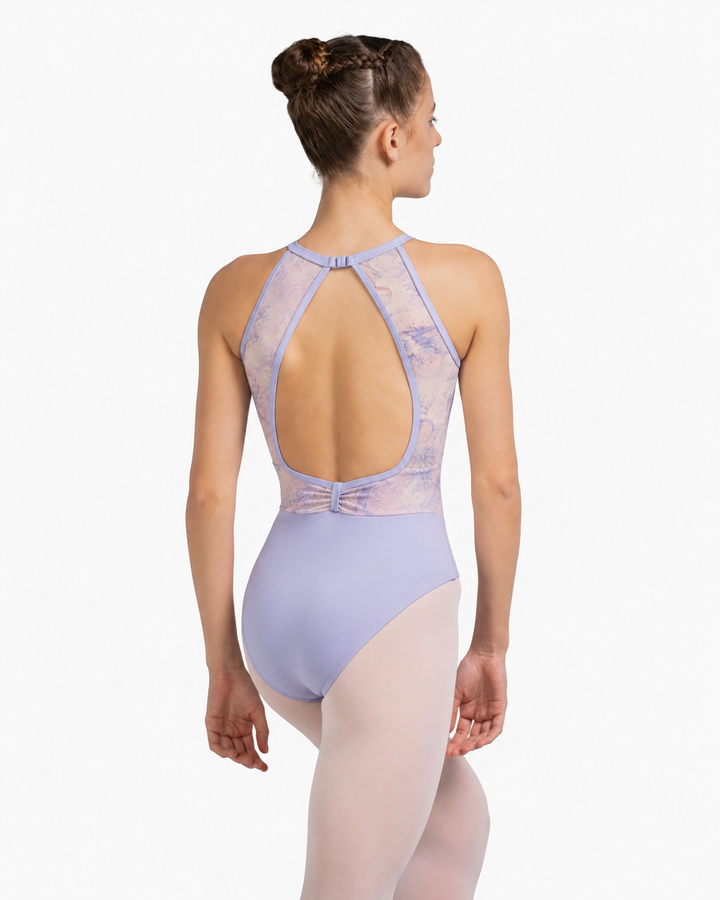 A woman modeling a lavender ballet leotard from the back, showing a high open back with sheer mesh straps and a bow detail at the waist, paired with light pink tights against a light gray background.