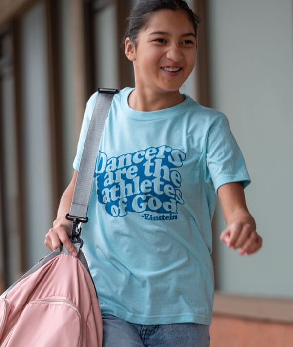 Person wearing a light blue t-shirt with text, Dancers are the athletes of God holding a pink bag indoors.