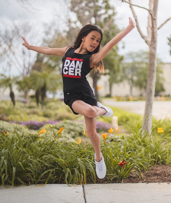 Young girl in a 'DANCER' tank top jumping outdoors in a park.
