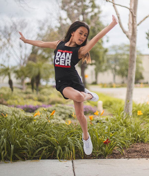Young girl in a 'DANCER' tank top jumping outdoors in a park.