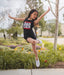 Young girl in a 'DANCER' tank top jumping outdoors in a park.