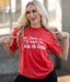 Woman wearing a red t-shirt with a humorous message, standing outdoors.