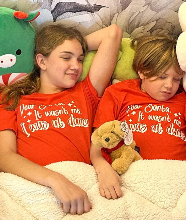 Two children wearing red shirts with humorous text, lying on a bed with stuffed animals.