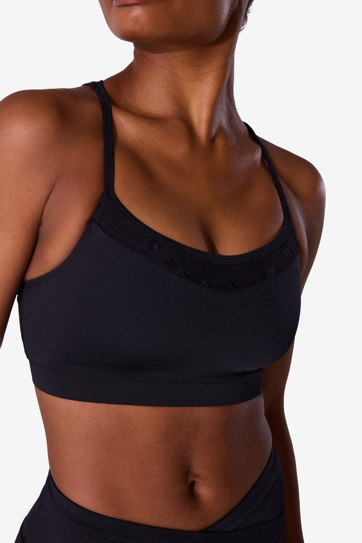 A close-up of a model wearing a black sports bra with delicate lace-up detailing around the scoop neckline, set against a plain light gray background.