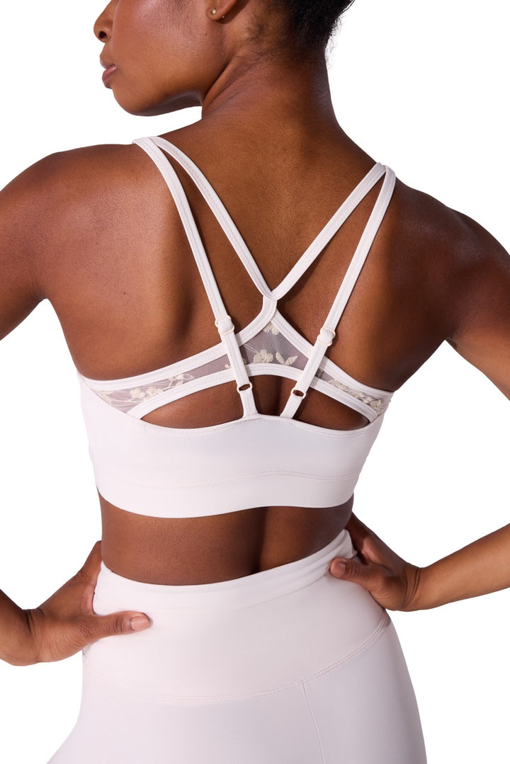 A close-up back view of a woman wearing a white sports bra with multiple crisscross lace-up straps and a sheer mesh panel at the upper back, paired with a white high-waisted athletic bottom against a plain white background.