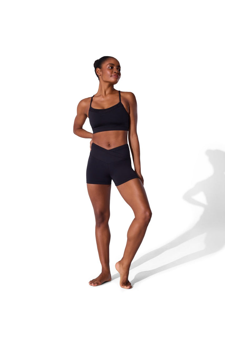 A woman modeling a matching black activewear set consisting of a sports bra and high-waisted shorts, standing against a plain white background with a soft shadow.