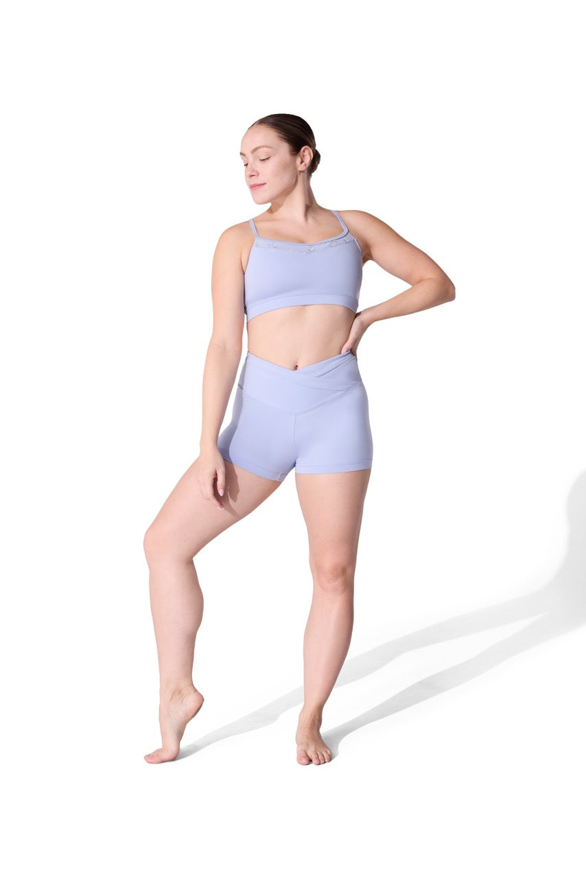 A woman modeling a light periwinkle blue two-piece activewear set consisting of a sports bra and high-waisted shorts, posed against a solid white background.