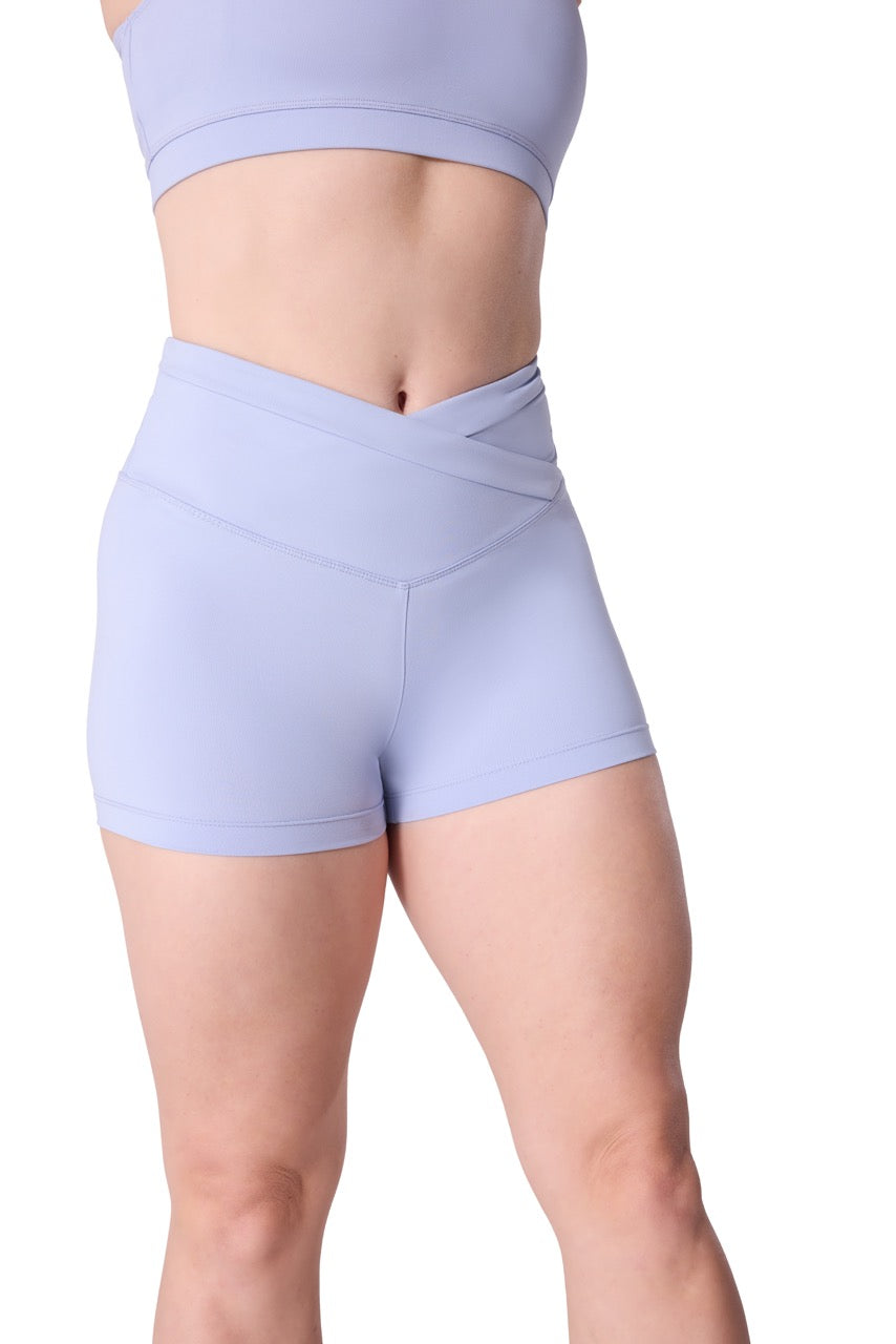 A close-up of a model wearing light periwinkle blue athletic shorts with a crossover waistband and a matching sports bra against a white background.