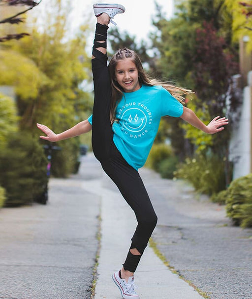 Person in a blue t-shirt and black leggings jumping on a sidewalk with trees in the background