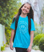 Young girl wearing a blue t-shirt with a logo and text, standing outdoors.