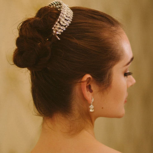 Woman with elegant hair bun and tiara, wearing a diamond earring, against a neutral background