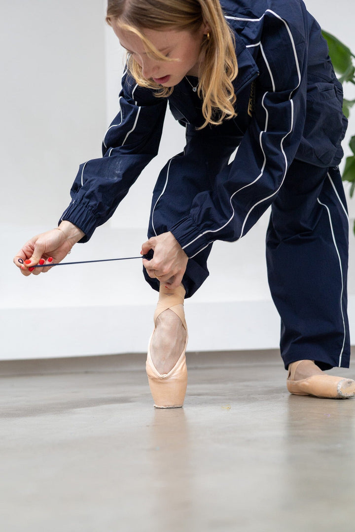 Person in navy outfit with white piping tying ballet shoes on a plain floor.
