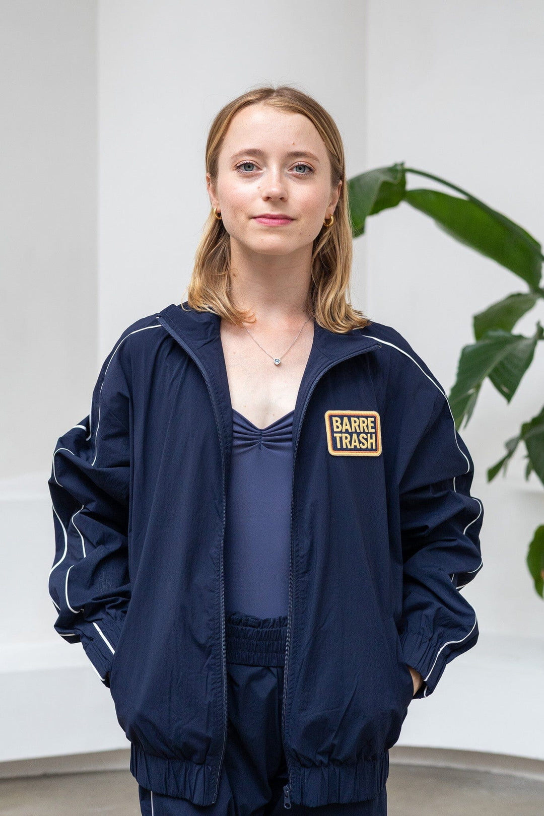 Person wearing a navy blue jacket with a patch, standing indoors with a plant in the background.