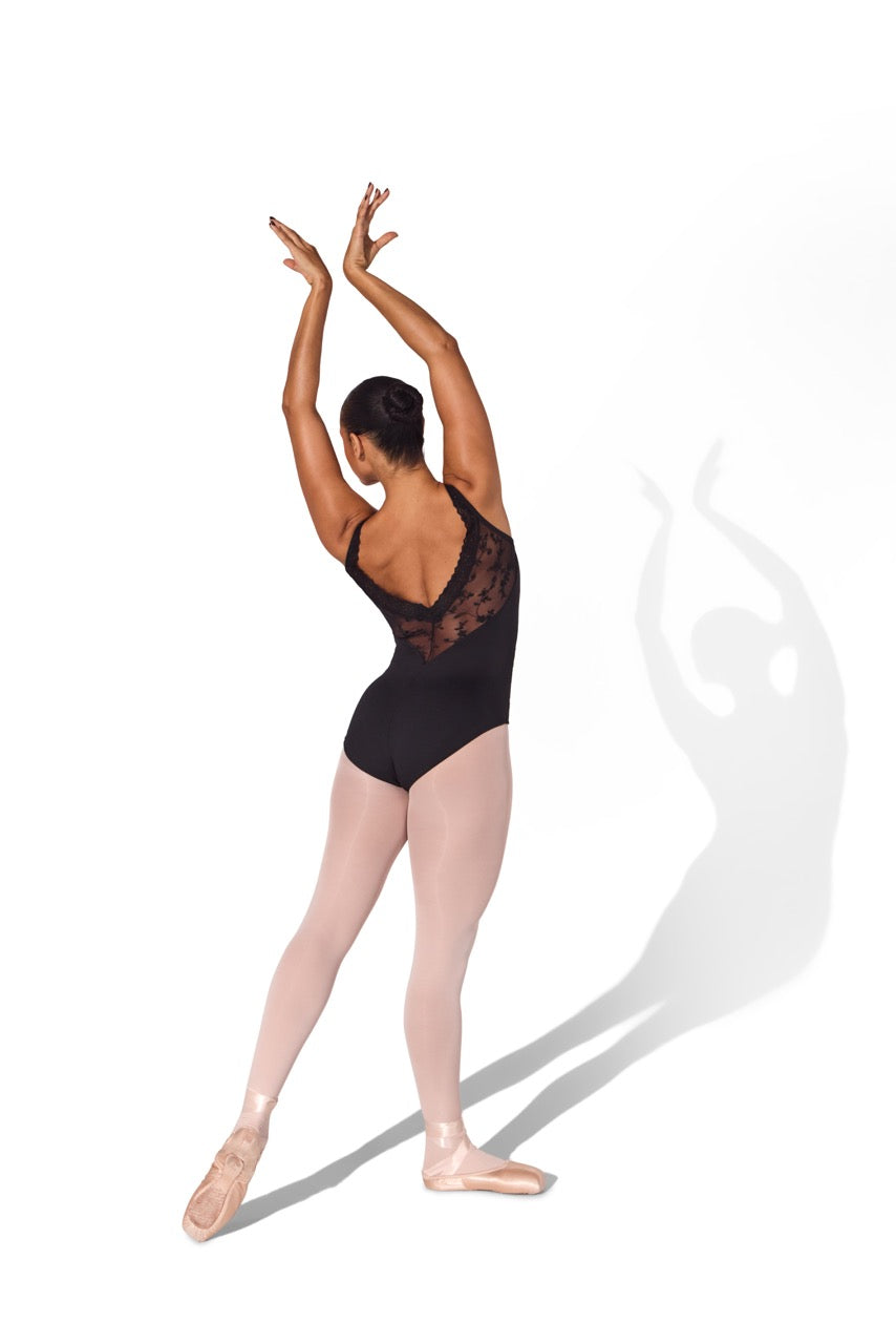 A woman in a black lace-back leotard and pink tights poses in a ballet position with her arms raised, photographed against a plain white background with a soft shadow.