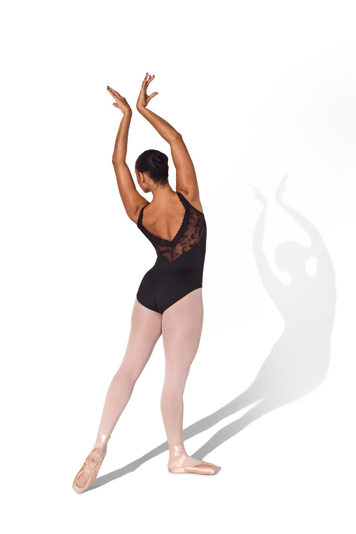 A woman in a black lace-back leotard and pink tights poses in a ballet position with her arms raised, photographed against a plain white background with a soft shadow.