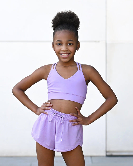 Young girl wearing a purple sports bra and shorts set against a white background
