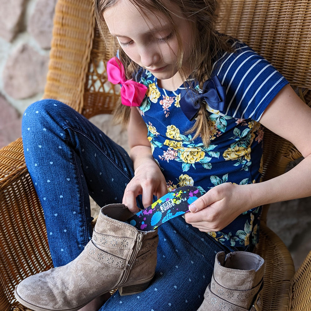 Young girl sitting on a wicker chair wearing a blue dress with floral patterns and polka dot pants, holding a colorful object.