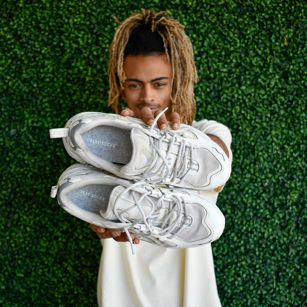 Person holding a pair of white sneakers against a green hedge background